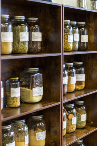 Wooden shelves with glass jars containing various herbs and spices, each labeled.